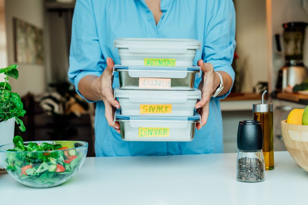 Person holding labeled meal prep containers for breakfast, lunch, snack, and dinner.