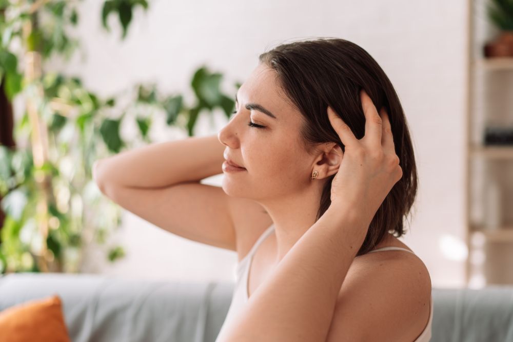 Woman with short hair enjoying a scalp massage, with her eyes closed in relaxation. The background shows indoor plants and a cozy living room setting, suggesting a peaceful, comfortable atmosphere.