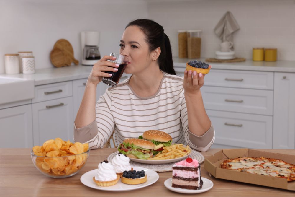 Woman sitting at a table full of fast food, holding a tart with blueberries while drinking a soda. The table includes items like chips, a hamburger with fries, desserts, and pizza, representing an assortment of unhealthy food choices.