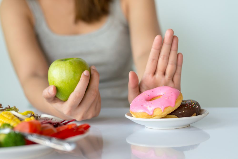 Person holding a green apple while rejecting donuts on a plate, symbolizing a healthy choice over sugary snacks. Represents a concept of healthy eating and lifestyle decisions.