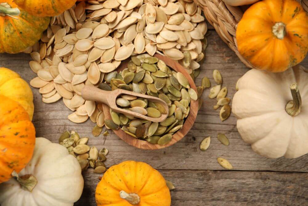 Pumpkin seeds in a wooden bowl with scattered seeds and pumpkins