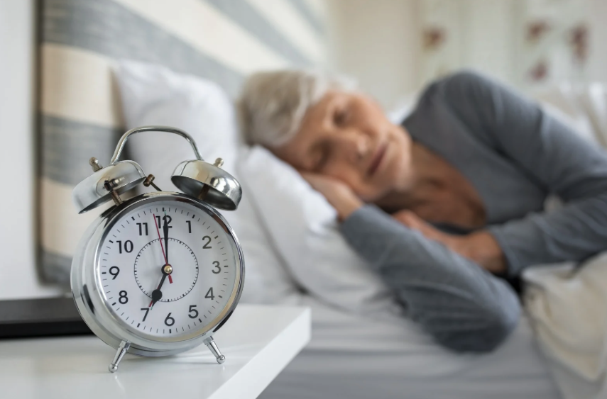 **Alt Text:**
An elderly woman sleeps peacefully in bed while a silver analog alarm clock shows the time as 6:53. The focus is on the clock in the foreground, symbolizing the importance of sleep and routine for health and wellness.
